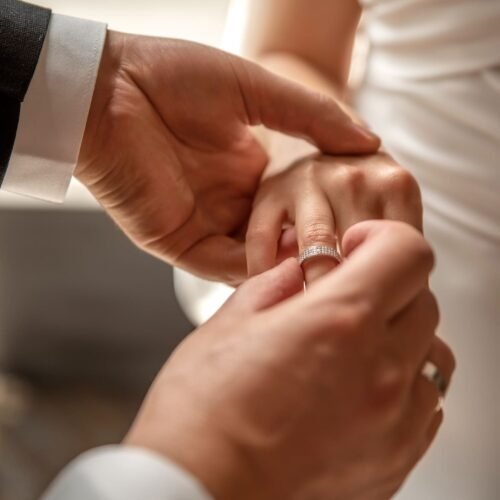 A groom puts on a diamond engagement ring on the bride's hand.